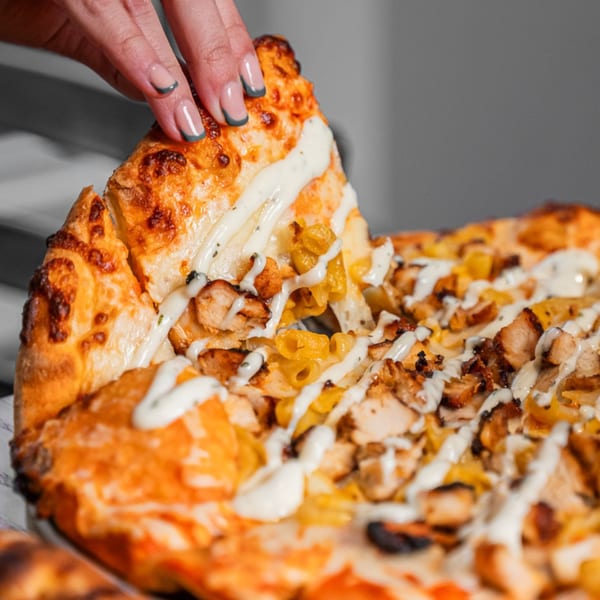 A hand with painted nails lifts a slice of chicken, pepper, and ranch pizza from a whole pie.