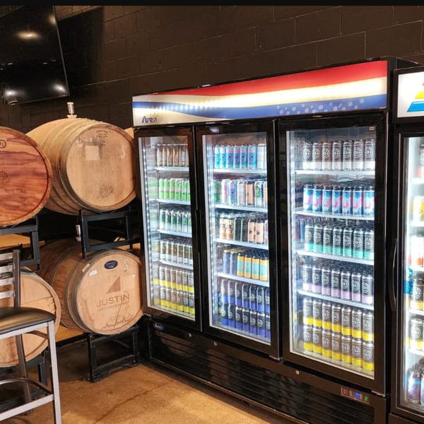 Three large refrigerators filled with various Loose Ends Brewery canned beers next to wooden barrels.