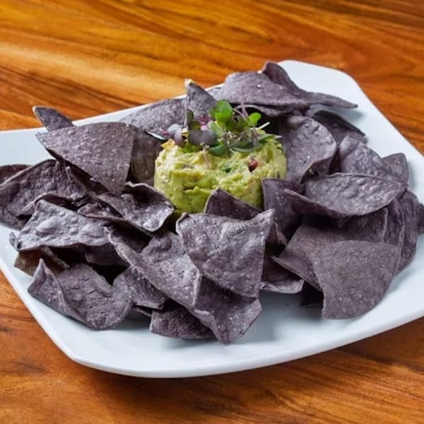 Blue corn tortilla chips surround a mound of guacamole, garnished with microgreens, on a white plate.
