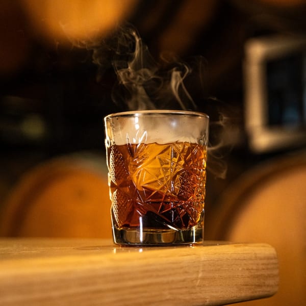 Steaming amber drink in a textured glass on a wooden surface, with blurred barrels in the background.