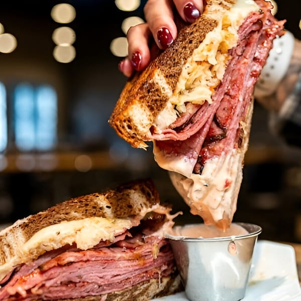 A hand holding up a reuben sandwich, dripping sauce into a metal cup at Loose Ends Brewery.